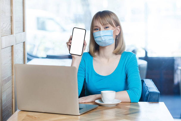 Portrait of young woman with surgical medical mask in blue t-shirt is sitting, looking at camera and showing her mobile copy space screen. Indoor working, medicine and health care concept. - Foto, Imagem