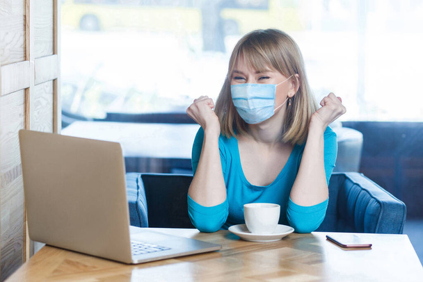 Yes. Portrait of happy young woman with surgical medical mask in blue t-shirt is sitting and working on laptop and celebrating her victory on work. Indoor working, medicine and health care concept. - Foto, Imagem