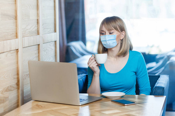 Portrait of young woman with surgical medical mask in blue t-shirt is sitting and working and looking at laptop screen and drinking coffee. Indoor distance working, medicine and health care concept. - Foto, afbeelding