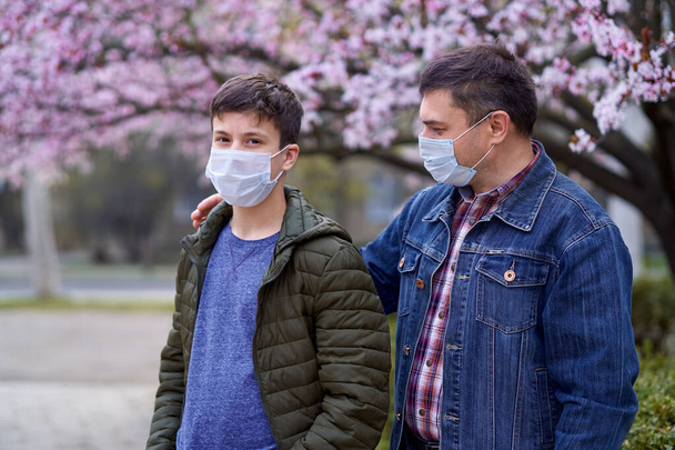 Vater und Sohn mit Gesichtsmaske sind in der Stadt draußen, blühende Bäume, Frühlingszeit, Blütezeit - Allergiekonzept und Gesundheitsschutz vor staubiger Luft - Foto, Bild