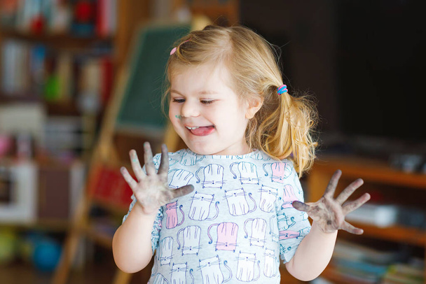 Мила чарівна дівчинка вчиться малювати з водними кольорами. Маленький малюк малює вдома, використовуючи барвисті пензлі. Здорова щаслива дитина з брудним обличчям і руками вдома або в дитячій
 - Фото, зображення