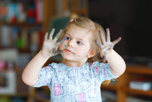 Cute adorable baby girl learning painting with water colors. Little toddler drawing at home, using colorful brushes. Healthy happy child with dirty face and hands at home or nursery - 写真・画像