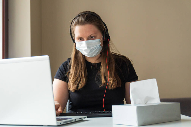 A girl in a medical mask works at home at the computer during the quarantine due to the coronavirus. Prevention of respiratory diseases. Remote work at home. - Photo, Image