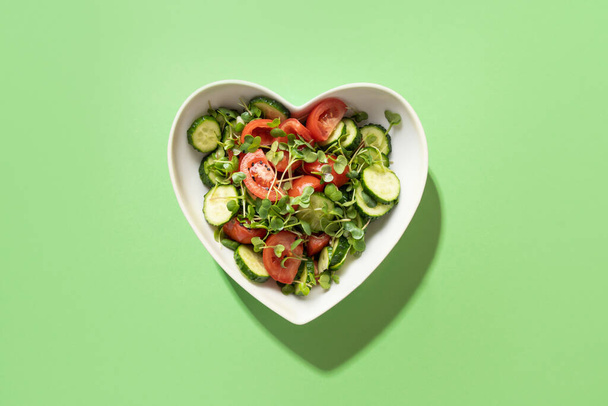 Fresh salad with tomato, cucumber, vegetables, microgreen radishes in plate shape of heart on green. View from above. Concept vegan and healthy eating. - Фото, изображение