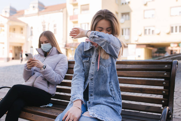 Mädchen in Masken. Coronavirus Thema. Frauen in der Stadt sitzen während der Quarantäne ohne Schutzmaske auf der Bank. Weibchen niesen und husten - Foto, Bild