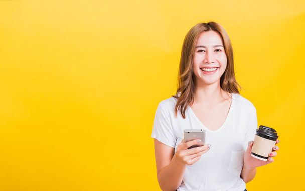 Portrait Asian Thai beautiful happy young woman standing smile, using mobile phone her holding coffee paper cup, looking to camera, studio shot isolated on yellow background, with copy space - Fotó, kép