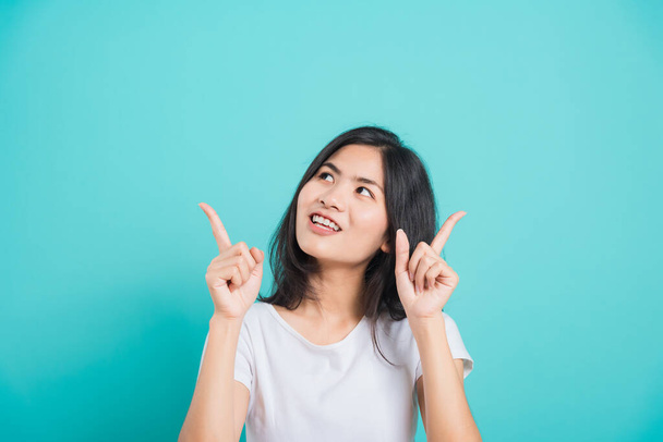 Portrait Asian beautiful young woman wearing white T-shirt standing smile white teeth, She pointing finger up and looking up, shoot photo in studio on blue background, There was copy space. - Photo, image