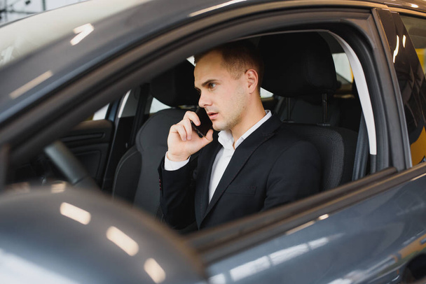 Young businessman sits in luxury car and talks on phone. He looks straight forward. Guy drives car. He holds one hand on steering wheel. It is sunny outside. - Foto, Bild