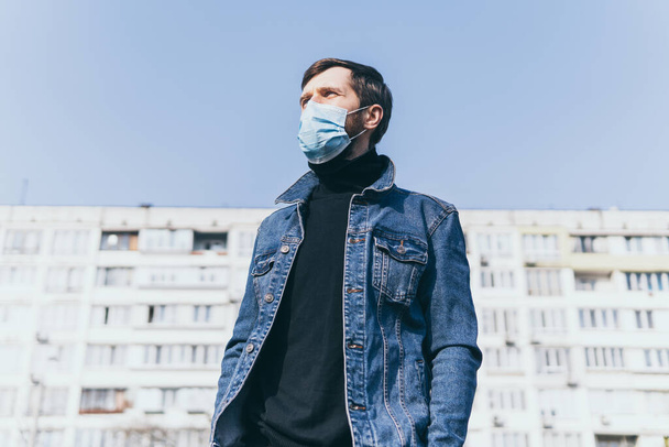 Concerned young man wearing medical mask outdoors with residential building on background. Corona virus quarantine conceptual image. - Photo, image