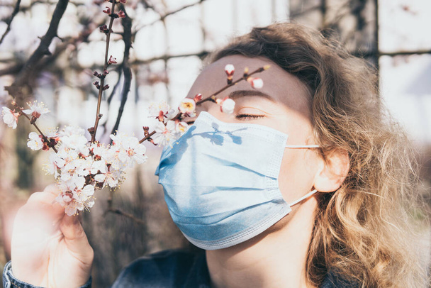 Young blonde woman smells cherry tree blossom through medical mask. Conceptual image of corona virus quarantine and allergy. - Photo, Image