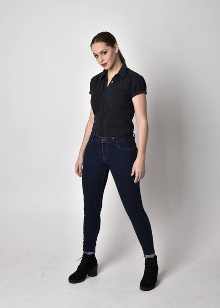 Portrait d'une jolie fille brune à queue de cheval, portant des bottes en jean et un chemisier noir. pose debout sur fond de studio
. - Photo, image