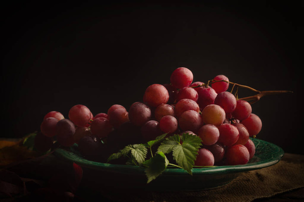 Świeży dojrzały różowy winogrono na vintage talerz, na starym drewnianym tle. Dark Moody Zdjęcie filtrowane w stylu światła Rembrandt. - Zdjęcie, obraz