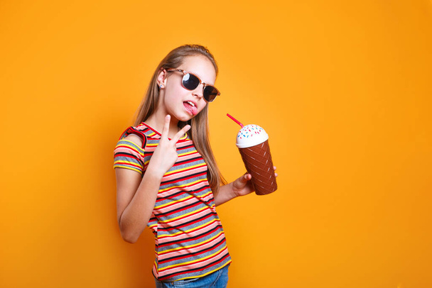 Cute caucasian young girl in t-shirt and sunglasses drinking milk cocktail on yellow studio background - Valokuva, kuva