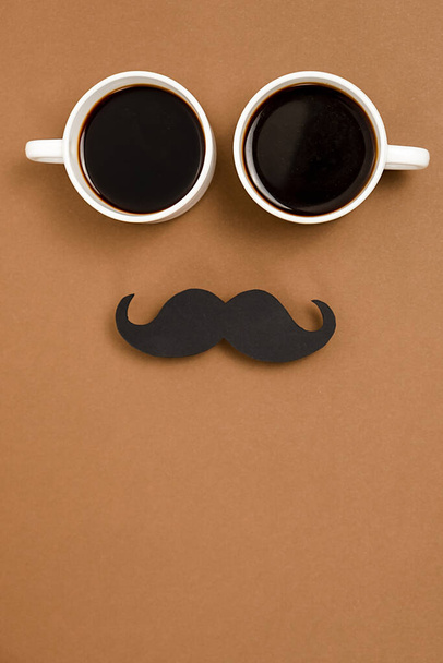 Vertical images of two cup coffee and retro stylish black paper photo booth props moustaches on brown background, space for text. Men health awareness month fathers day masculinity concept. - Foto, imagen