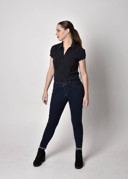 Portrait of a pretty brunette girl with ponytail, wearing jeans boots and a black blouse.  full length standing pose on a studio background. - Fotografie, Obrázek