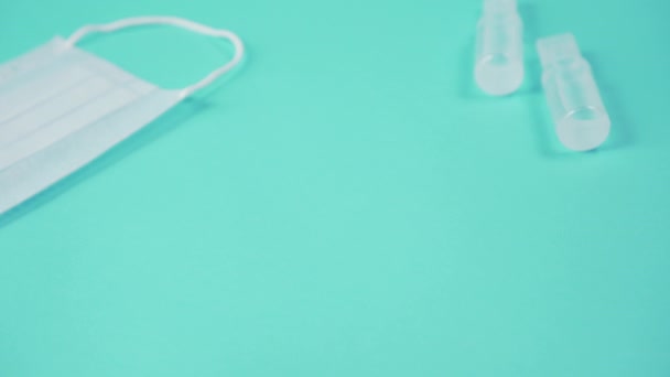 mão em luvas cirúrgicas coloca um pacote de ampolas de vidro com uma vacina em uma superfície azul ao lado de uma máscara protetora médica
 - Filmagem, Vídeo