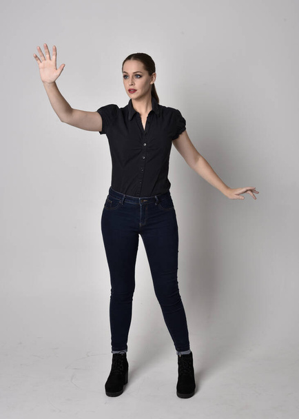 Портрет гарненької брюнетки з хвостом у джинсах та чорною блузкою. повнометражна стояча поза на студійному фоні
. - Фото, зображення