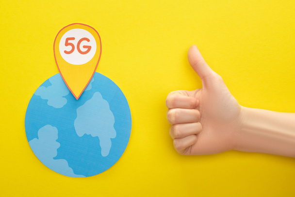 top view of globe and 5g lettering in location mark near thumb up on yellow background - Photo, Image