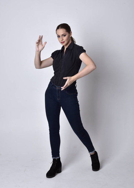 Portrait of a pretty brunette girl with ponytail, wearing jeans boots and a black blouse.  full length standing pose on a studio background. - 写真・画像