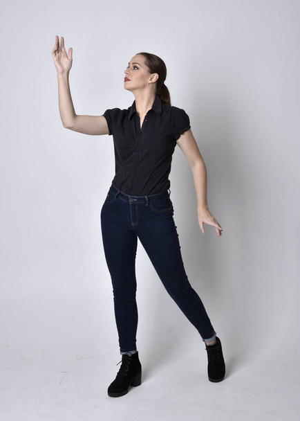 Portrait of a pretty brunette girl with ponytail, wearing jeans boots and a black blouse.  full length standing pose on a studio background. - Фото, зображення