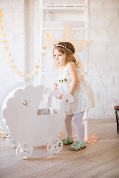 Little cute girl in a white dress plays with a white peg for dolls  in a bright children's room, decorated with toys and garlands. - Foto, Imagen
