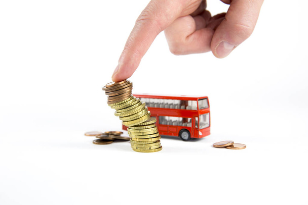 Man vinger duw over de stapel munten, rode miniatuur bus op de achtergrond, geïsoleerd op witte achtergrond. Kosten in het vervoerssysteem. - Foto, afbeelding