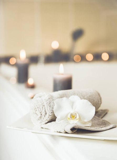 Pequeña toalla enrollada con flor de orquídea blanca en bandeja de cerámica blanca, velas ardientes en el fondo en el cuarto de baño. concepto de lavandería limpia en seco fresco
. - Foto, imagen