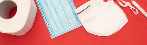 top view of white toilet paper, safety and medical masks and thermometer on red background, panoramic shot - Photo, Image
