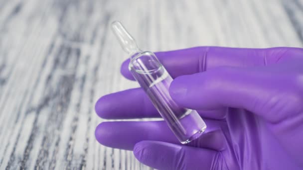 Medical glass ampoule in hand in blue protective surgical gloves on wooden background - Πλάνα, βίντεο