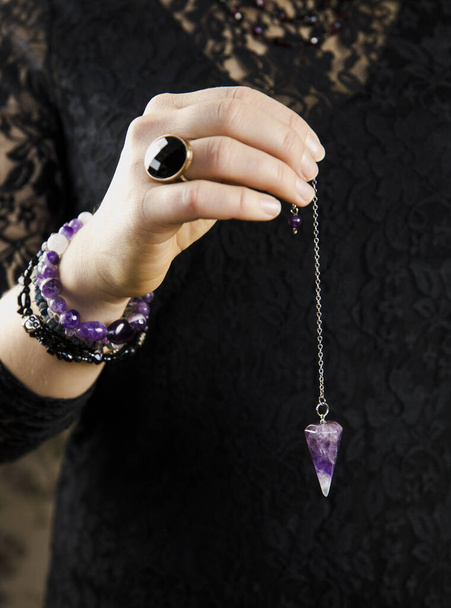 Close up of woman wearing black gothic clothing, hand holding and using amethyst crystal pendulum on silver chain fortune telling pendulum in hand. Mystical psychic dark witch concept.  - Photo, Image