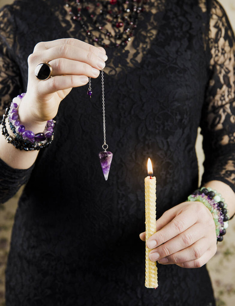 Close up of elegant woman wearing black gothic clothing, hand holding and using amethyst crystal pendulum on silver chain fortune telling pendulum in hand.     - Photo, Image