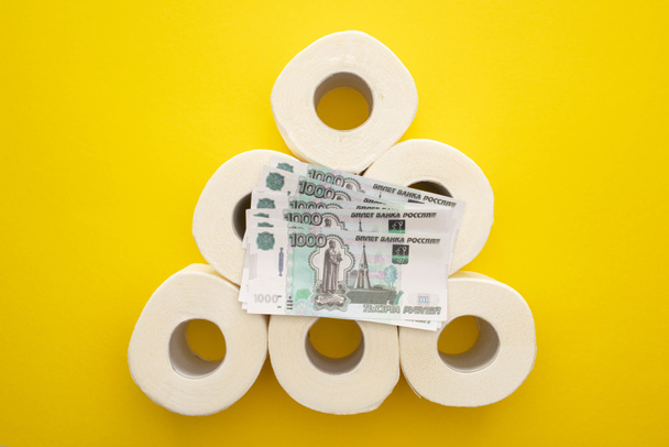 Ansicht von oben weiße Toilettenpapierrollen in Pyramide mit russischem Geld auf gelbem Hintergrund angeordnet - Foto, Bild