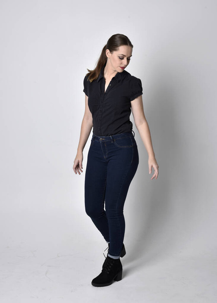 Portrait of a pretty brunette girl with ponytail, wearing jeans boots and a black blouse.  full length standing pose on a studio background. - Zdjęcie, obraz