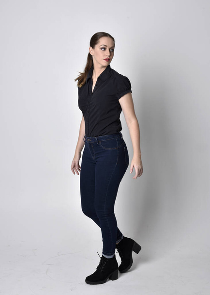 Portret ładnej brunetki z kucykiem, w dżinsach i czarnej bluzce. pełna długość stojąca poza na tle studia. - Zdjęcie, obraz