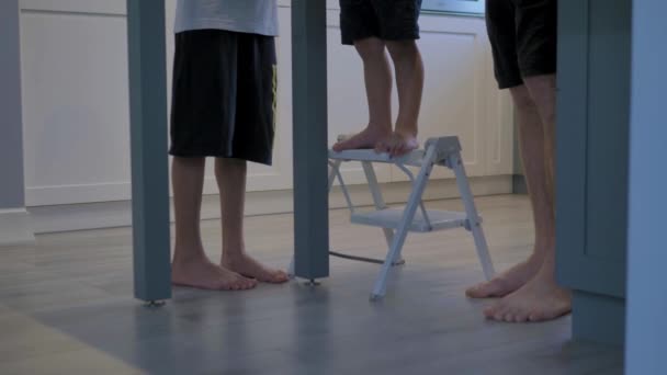 The legs of two sons of different ages, and a father, are standing on the wooden floor next to the table. They are doing something. - Filmati, video