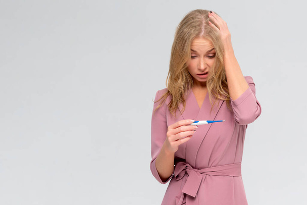 Schockierte kranke Frau blickt auf Thermometer isoliert auf weißem Hintergrund. Symptome des Covid-19-Coronavirus - Foto, Bild