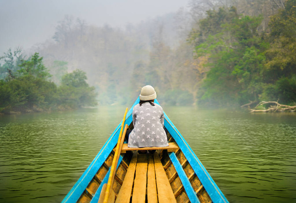 Невеликий дерев'яний човен, що плаває в дикій тропічній річці в зелених джунглях з жінкою, сидячи перед човном і спокійно дивлячись на пейзажі. Блакитне каное досліджує природу
. - Фото, зображення