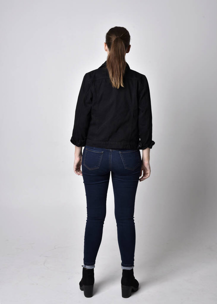 Portrait of a pretty brunette girl with ponytail, wearing jeans boots and a black blouse.  full length standing pose, with back to the camera on a studio background. - Photo, image