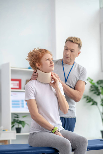 Male physiotherapist examining neck fixator of female patient sitting on couch - Φωτογραφία, εικόνα