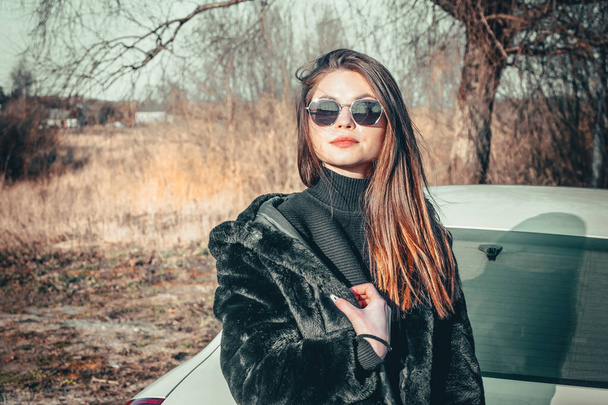 A brunette girl walks in the open air, dressed in a black fur coat and black sunglasses. - Foto, afbeelding