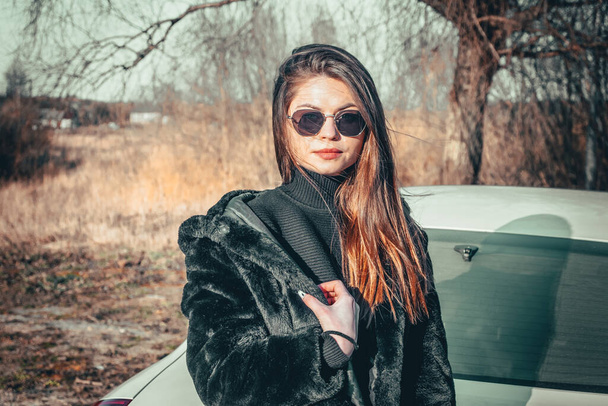 A brunette girl walks in the open air, dressed in a black fur coat and black sunglasses. - Foto, afbeelding