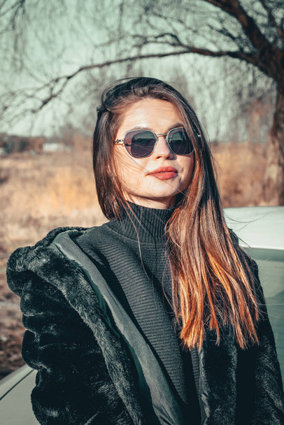 A brunette girl walks in the open air, dressed in a black fur coat and black sunglasses. - Valokuva, kuva