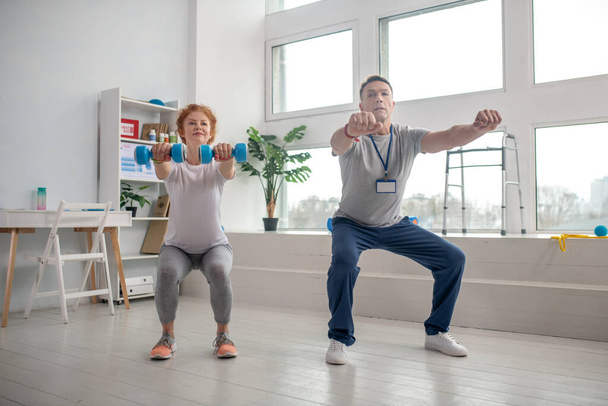 Female patient holding dumbbells and squatting together with male physiotherapist - Valokuva, kuva