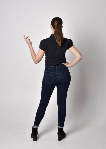 Portrait of a pretty brunette girl with ponytail, wearing jeans boots and a black blouse.  full length standing pose, with back to the camera on a studio background. - Photo, image