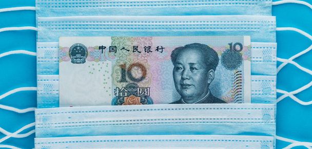 COVID-19 coronavirus in China. Stijging van de prijs van medische maskers. Medische masker en bankbiljet van 10 yuan op blauwe achtergrond, plat lay. Export van medische maskers uit China. Bovenaanzicht, close-up. - Foto, afbeelding