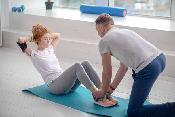 Female patient doing sit-ups on mat, male physiotherapist helping her by holding her ankles - 写真・画像