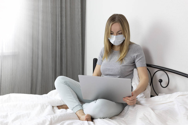 Tıbbi maskeli, gri tişörtlü ve beyaz bir yatakta dizüstü bilgisayarlı rahat pantolonlu genç hasta bir kadın. Coronavirüs salgını yüzünden evden çalışıyor. salgın COVID-19 - Fotoğraf, Görsel