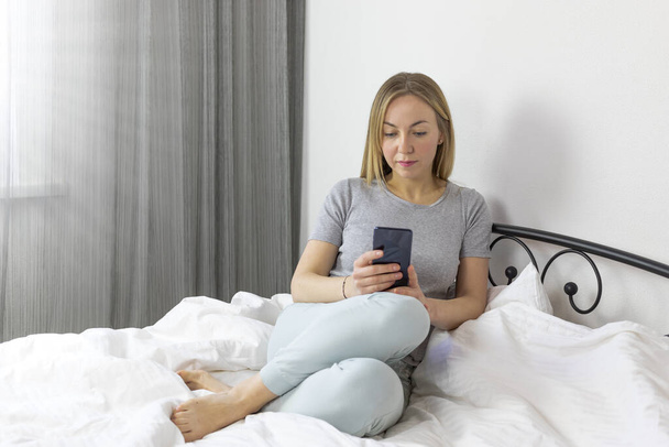 Young pretty european blonde woman working and studying from home . in bed with smartphone. she is in light, white, comfortable home clothes. - Photo, Image