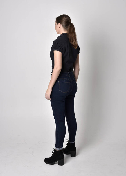 Portrait of a pretty brunette girl with ponytail, wearing jeans boots and a black blouse.  full length standing pose, with back to the camera on a studio background. - Foto, Imagem