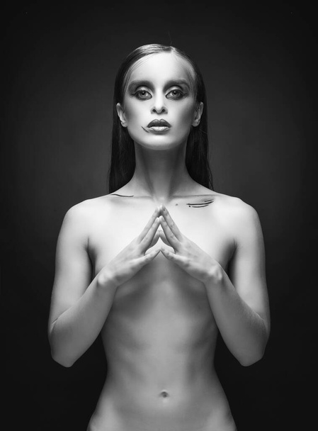 A beautiful slim topless girl with green avant-garde makeup sensually poses covering her breast with her hands, on a dark background.  Artistic, fashionable, advertising monochrome design - Foto, afbeelding